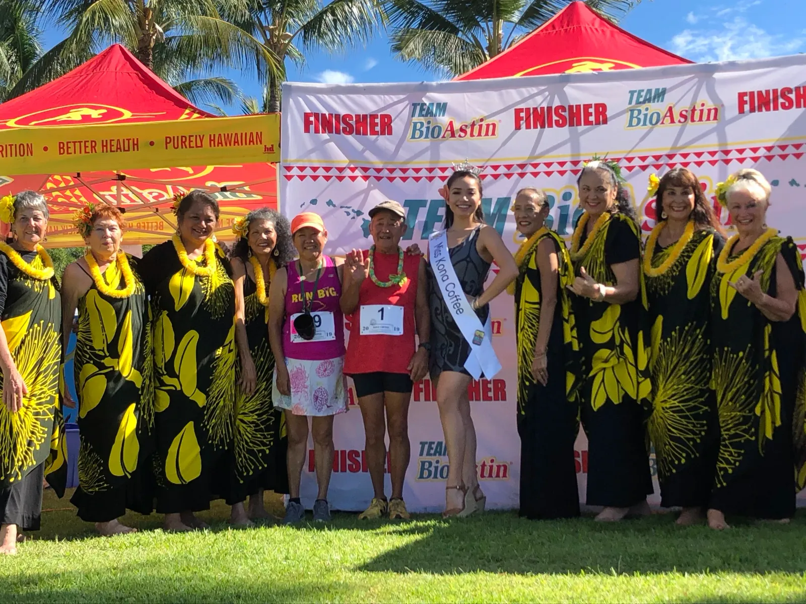 A group of people, including finishers and a beauty queen, pose for a photo at the Kona Coffee Half Marathon finish line, with traditional Hawaiian dancers in the foreground.