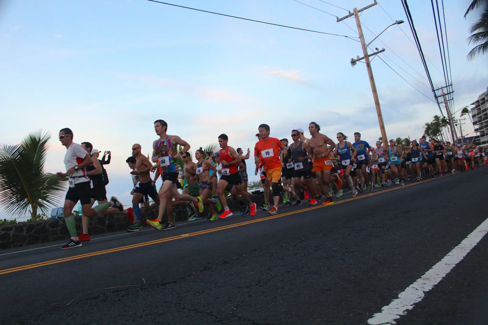 A group of runners are starting a race on a road with palm trees and buildings in the background.