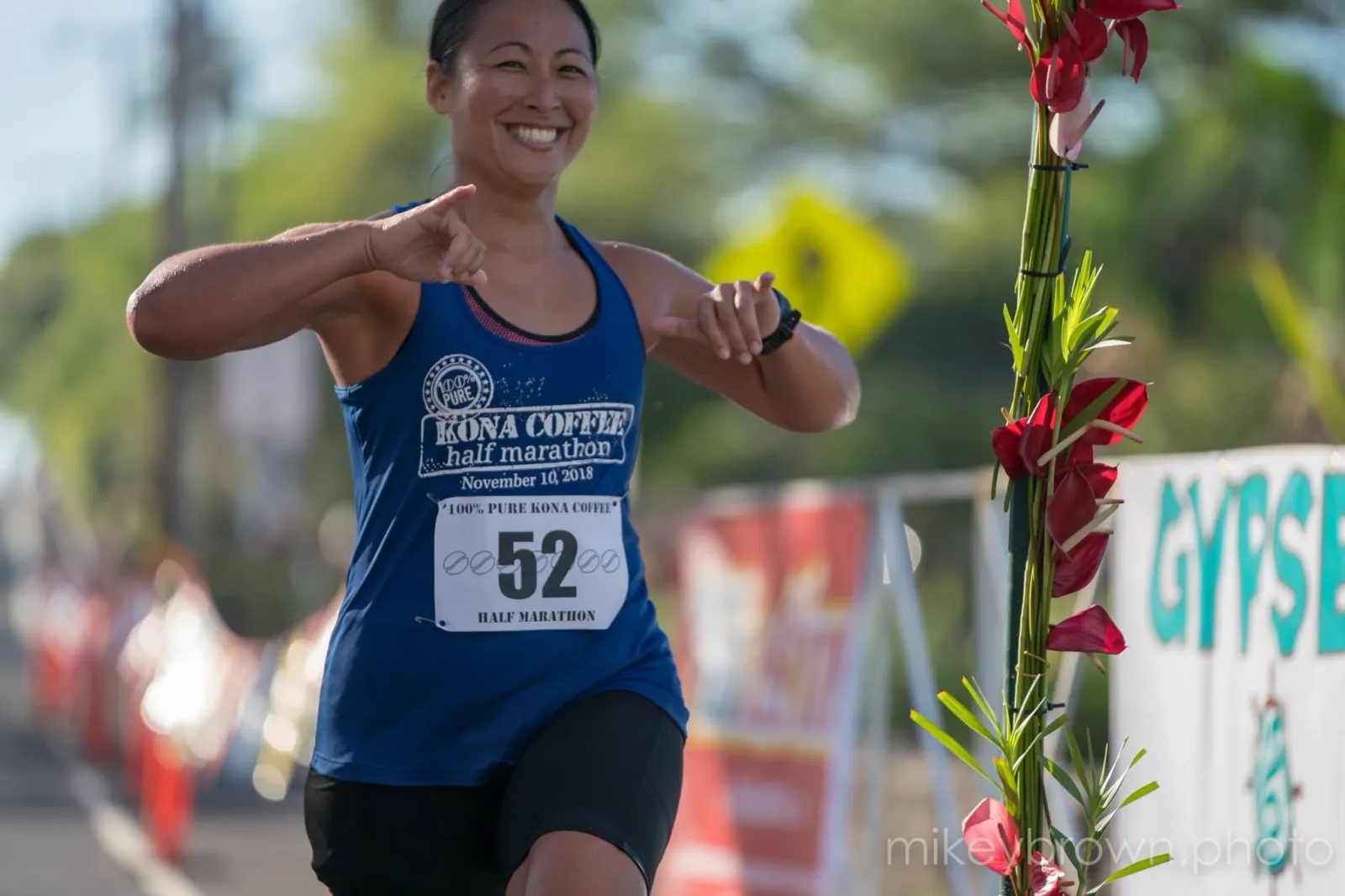A female runner smiles as she runs, wearing a blue race bib with the number 52 and the text 'Kona Coffee Half Marathon'.
