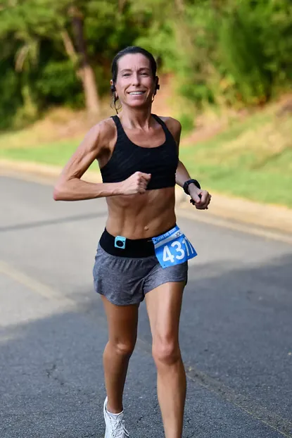 A female runner with bib number 431 smiles as she runs on a road with trees in the background.