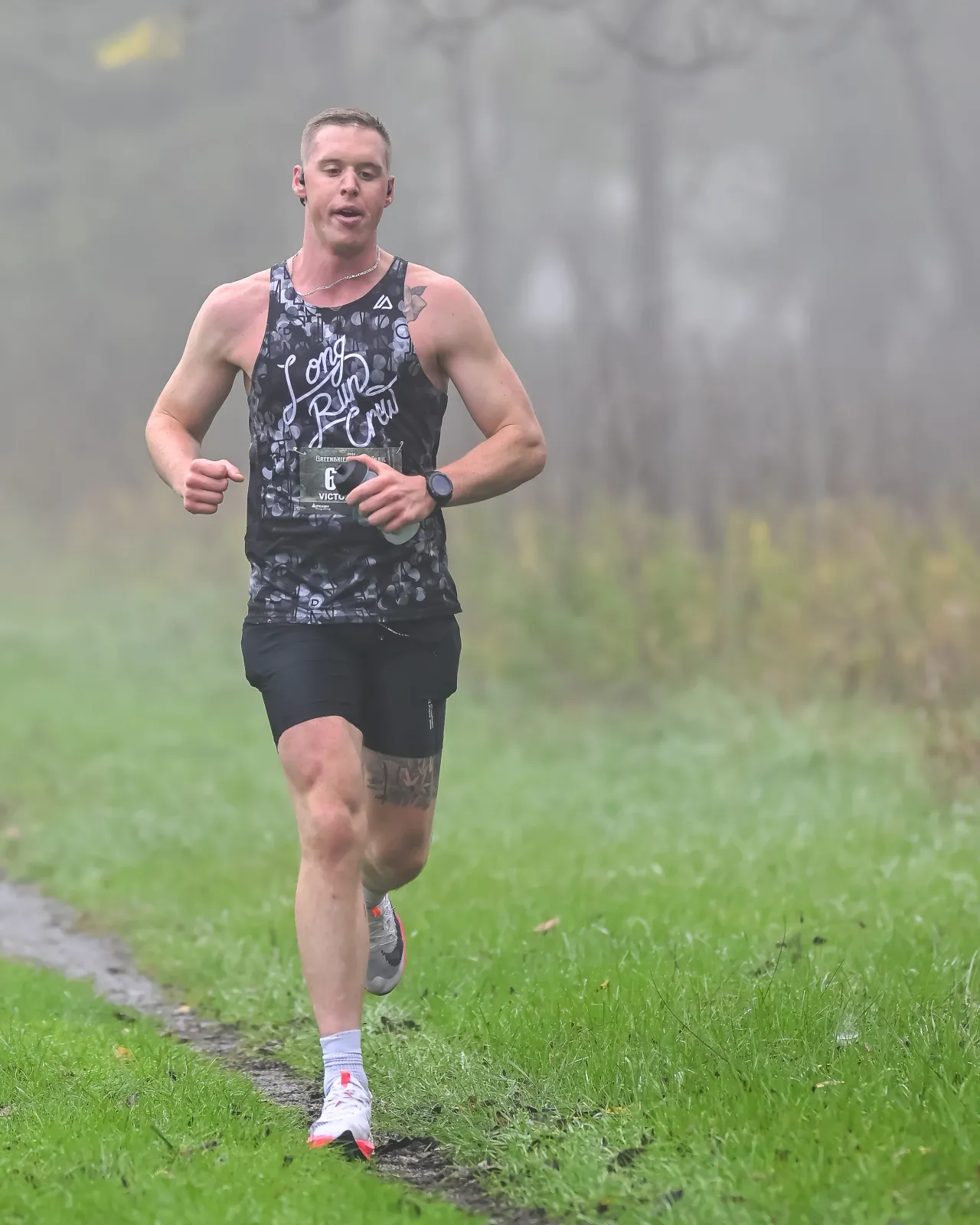 A male runner with a bib number 6 runs on a muddy trail with grass on the sides, wearing a black tank top and shorts, with fog in the background.
