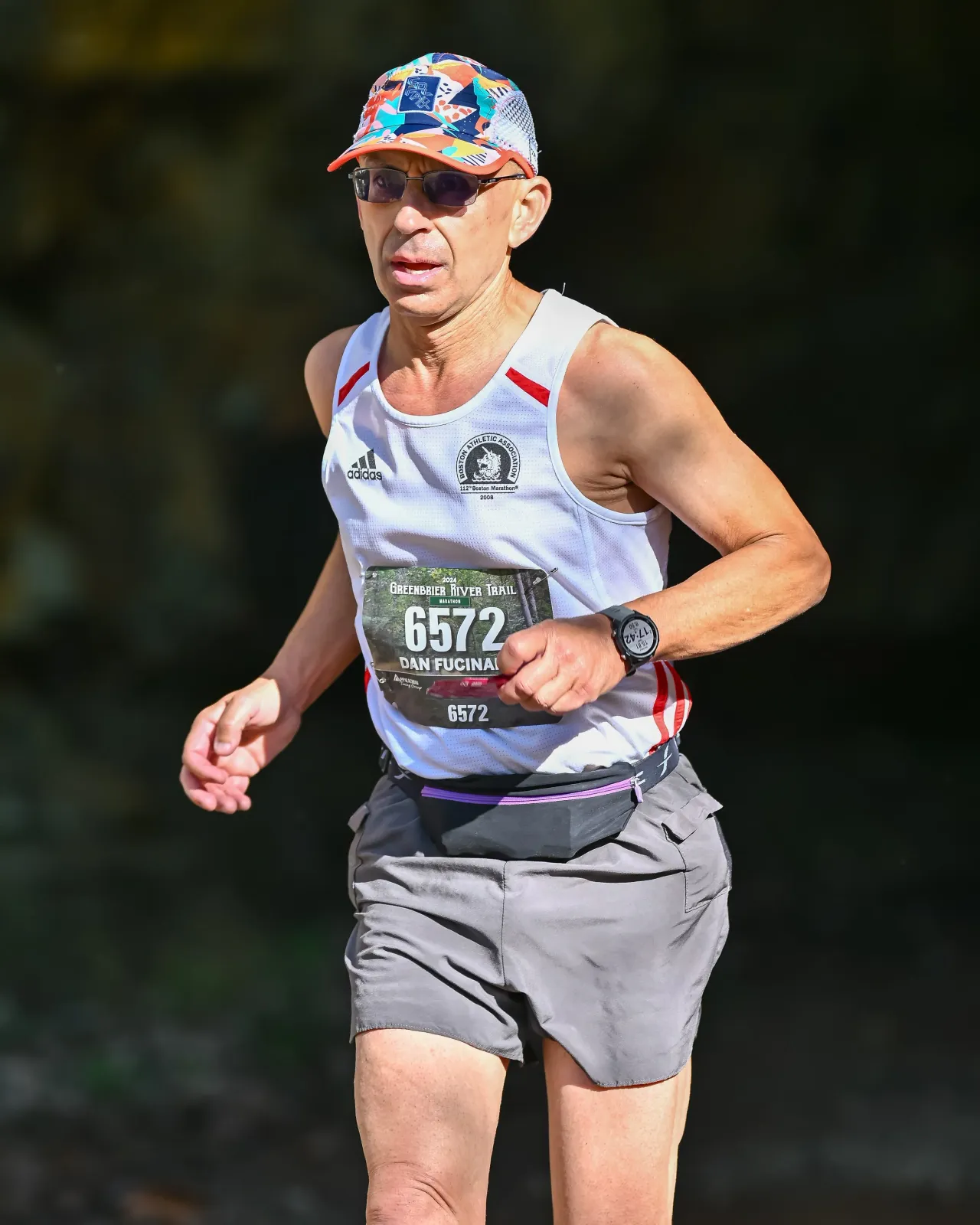 A male runner wearing a white tank top, grey shorts, sunglasses, and a colorful hat runs on a trail. He has bib number 6572 and a fanny pack.