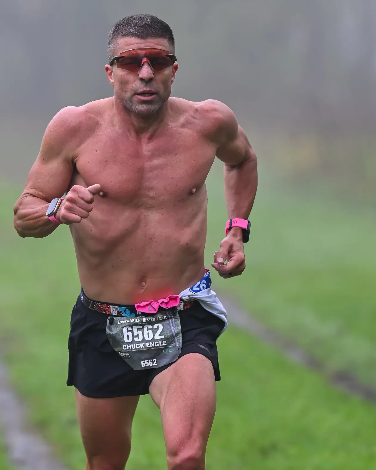 A shirtless male runner with sunglasses and bib number 6562 running on a trail.
