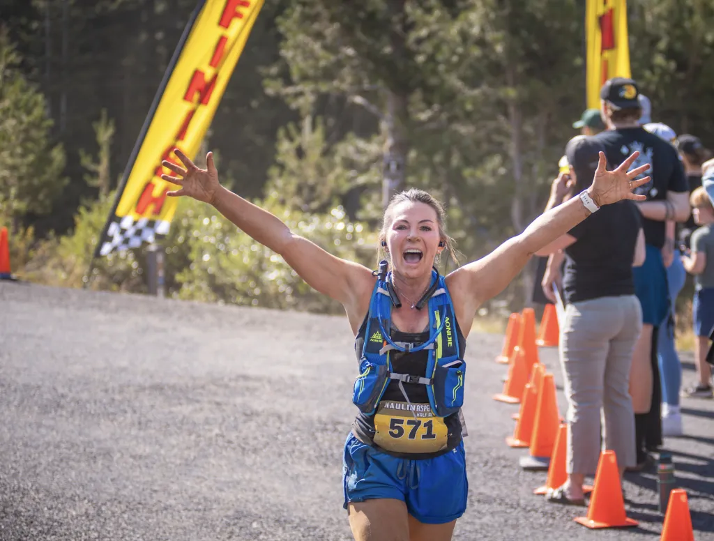 A female runner crosses the finish line with her arms raised in triumph, a look of exhilaration on her face. A 'FINISH' banner is visible in the background.