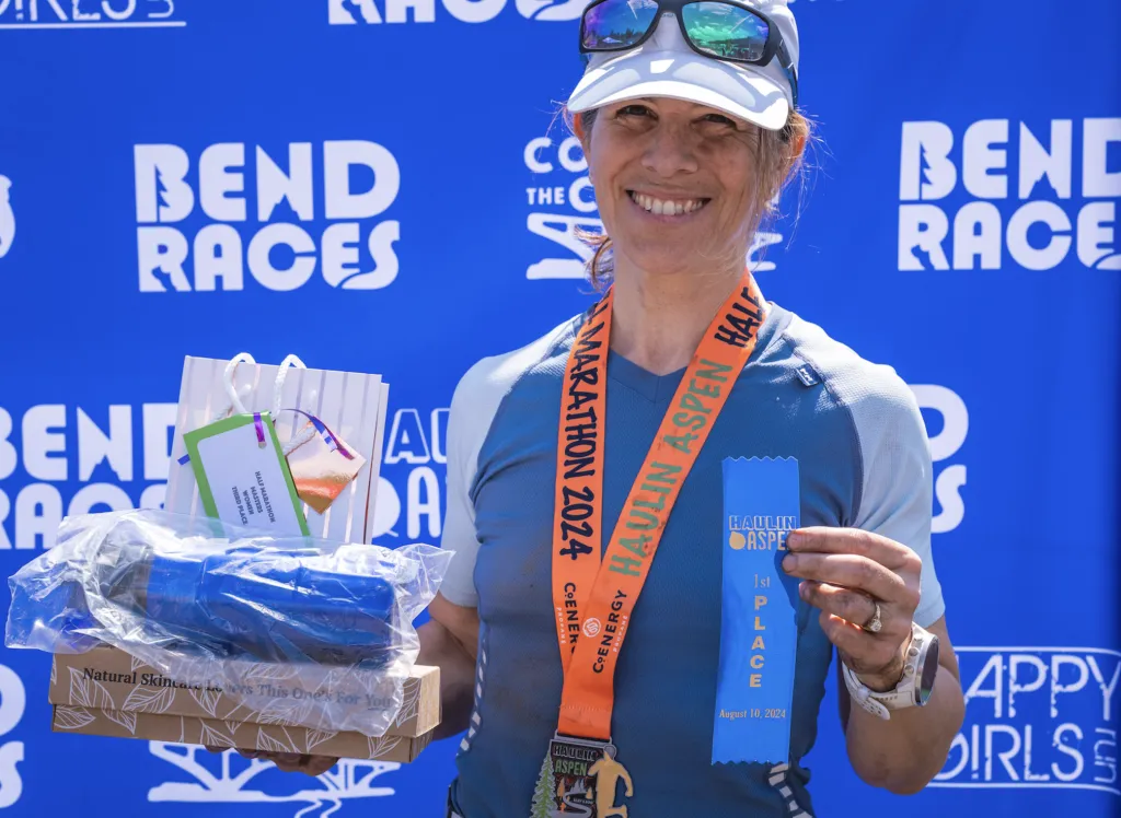 A smiling female runner holds a medal and a blue ribbon that says '1st PLACE' for the Haulin' Aspen Half Marathon on August 10, 2024. She is also holding a gift bag and a box of prizes.