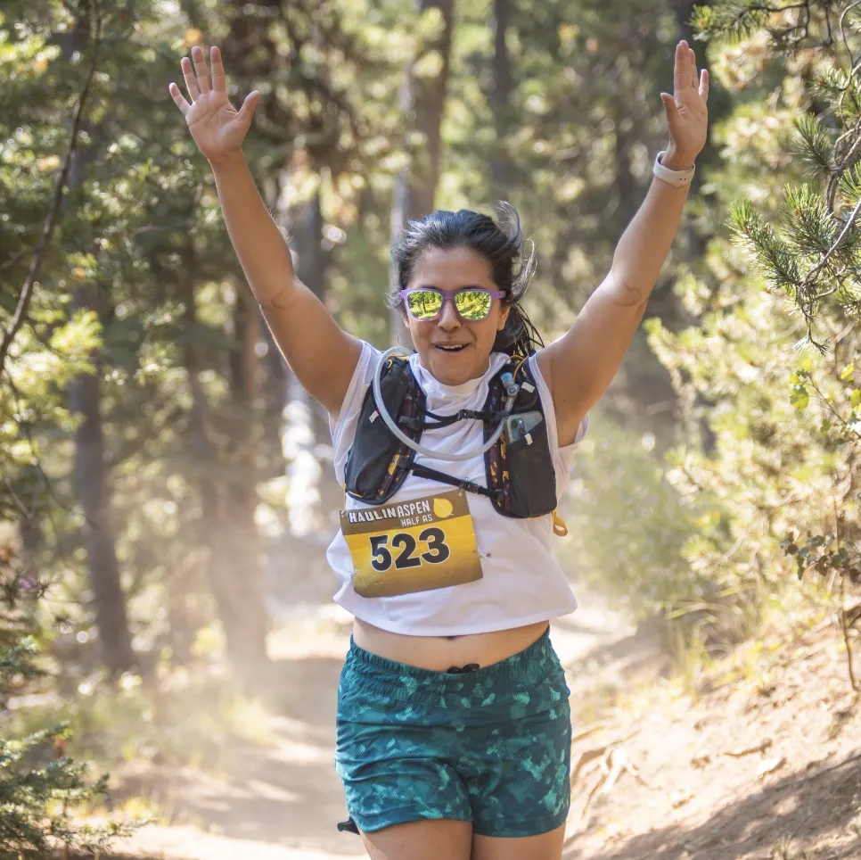 A female runner with her arms raised in celebration crosses the finish line on a wooded trail during the Haulin' Aspen Half Marathon.
