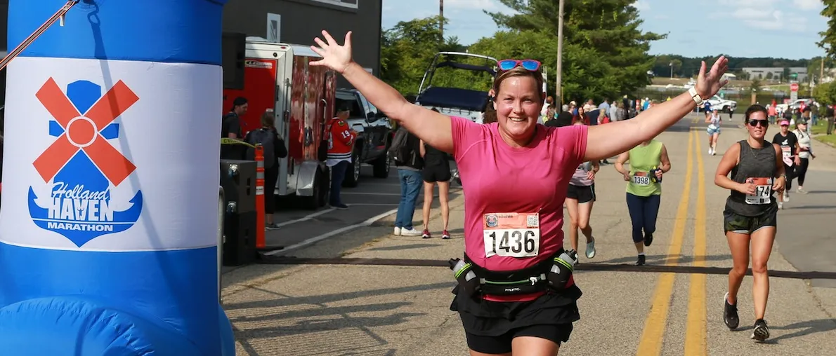 A female runner with arms outstretched in celebration crosses the finish line of the Holland Haven Marathon, with other runners and an inflatable arch in the background.