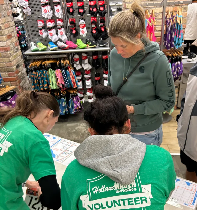 Volunteers wearing green shirts with 'Holland Haven Marathon VOLUNTEER' printed on them are looking at a map on a table.