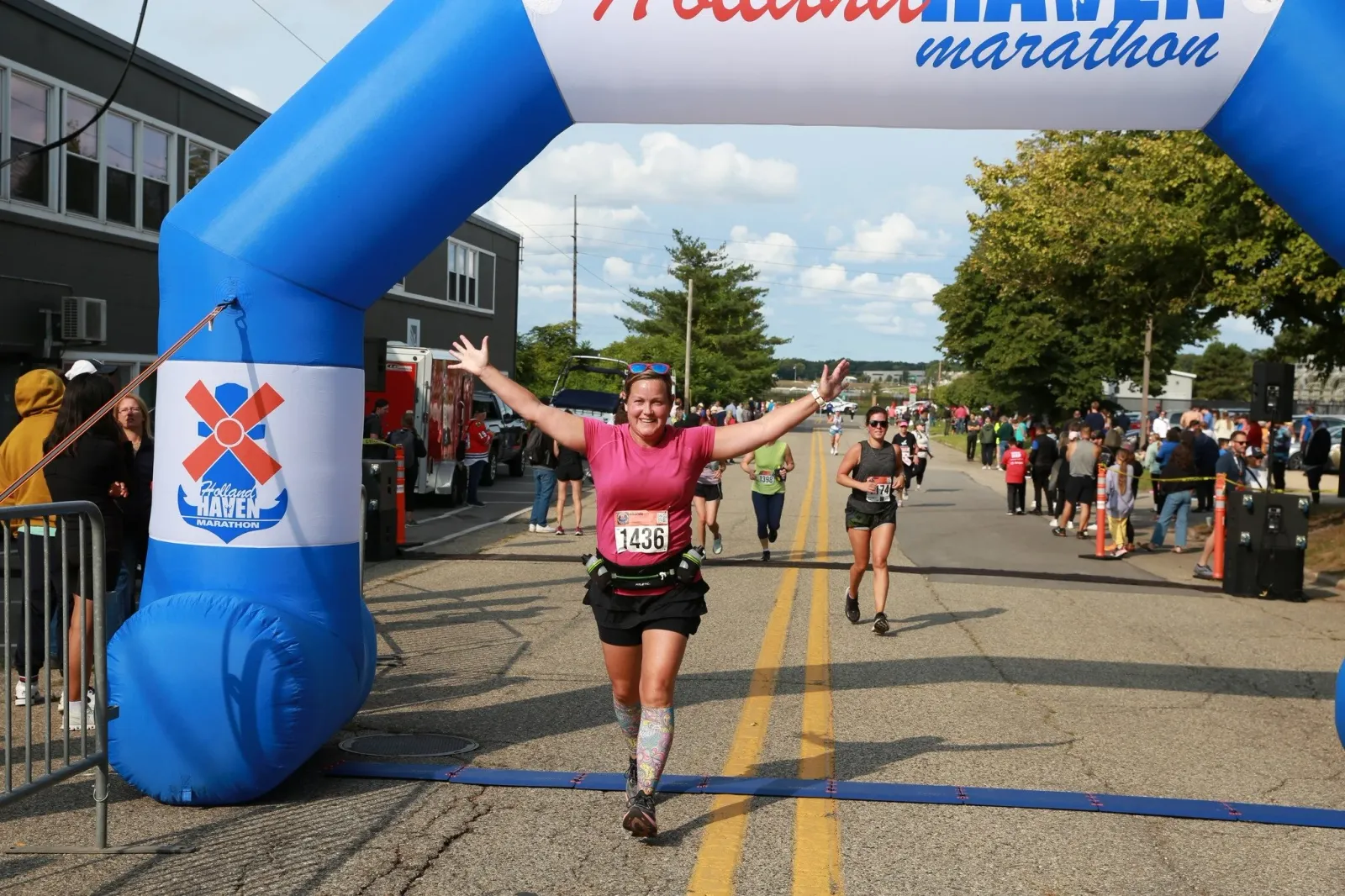 A female runner with arms outstretched in celebration crosses the finish line under a blue inflatable arch at the Holland Haven Marathon.