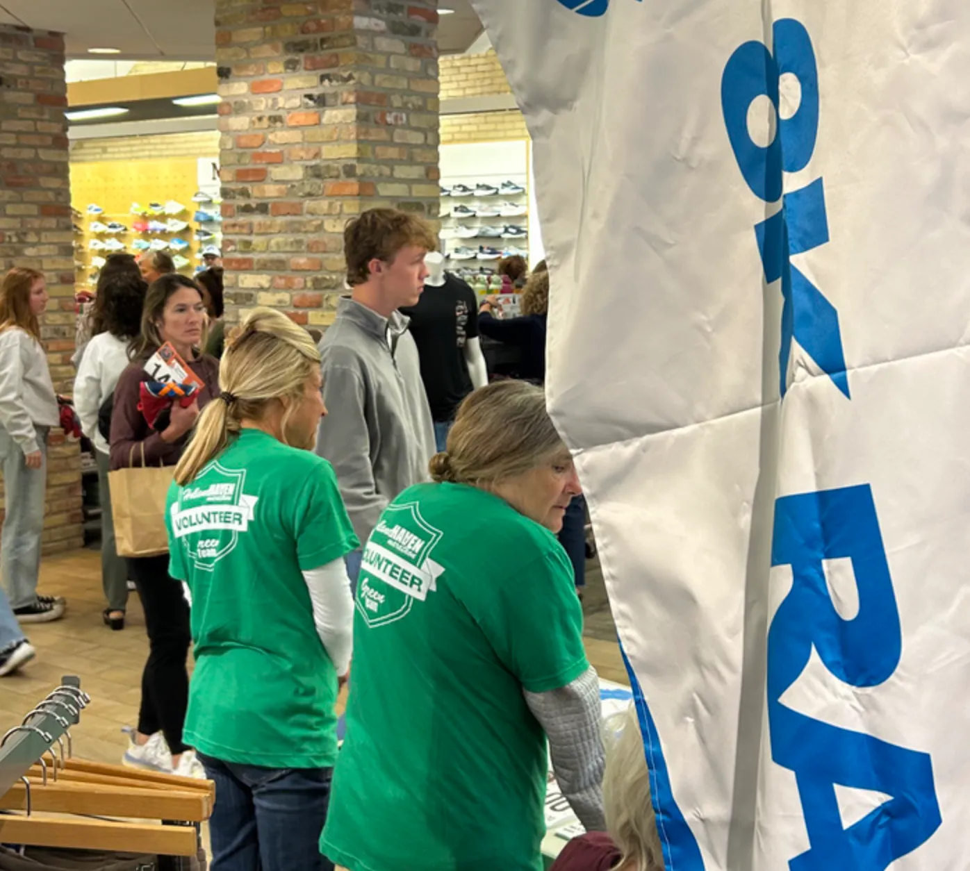 Volunteers wearing green shirts with 'Holland Haven Volunteer' printed on them, standing near a large white banner with blue lettering, inside a building with shelves of shoes in the background.