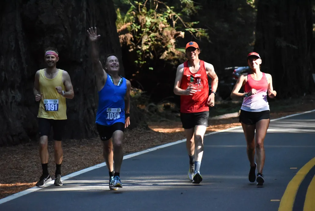 Runners participating in the Humboldt Redwoods Marathon, Half Marathon, or 5k, running on a paved road surrounded by redwood trees. One runner waves to the camera.