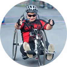 A wheelchair athlete wearing a helmet and sunglasses races forward on a paved road.
