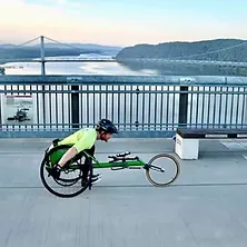Wheelchair athlete racing across a bridge with a river and hills in the background at sunset.