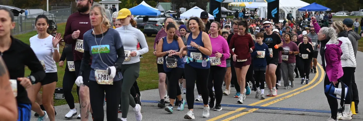 A large group of runners are at the start line of the Rhinebeck Fall Foliage Half Marathon & 5K, with a finish line arch in the background.