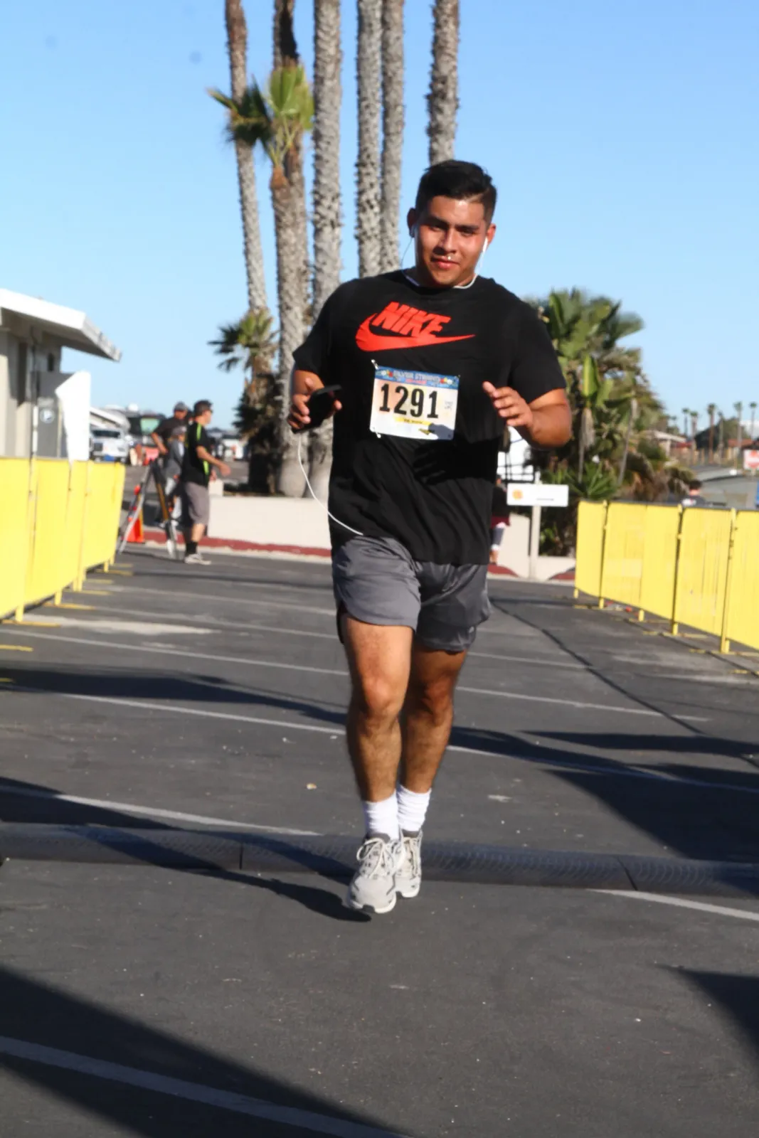 A male runner wearing a black Nike t-shirt and grey shorts runs towards the camera, wearing a bib with the number 1291.