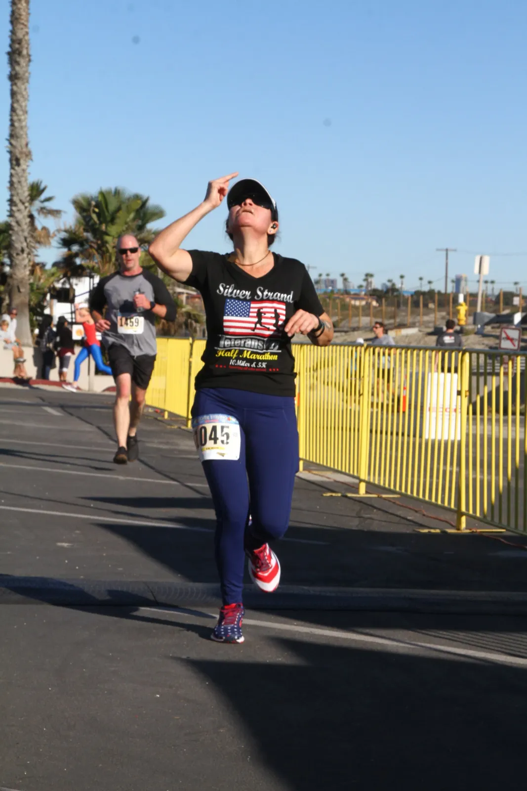 A female runner wearing a black t-shirt with 'Silver Strand Veterans Half Marathon' and an American flag graphic, running on a road with a yellow barrier to her right.