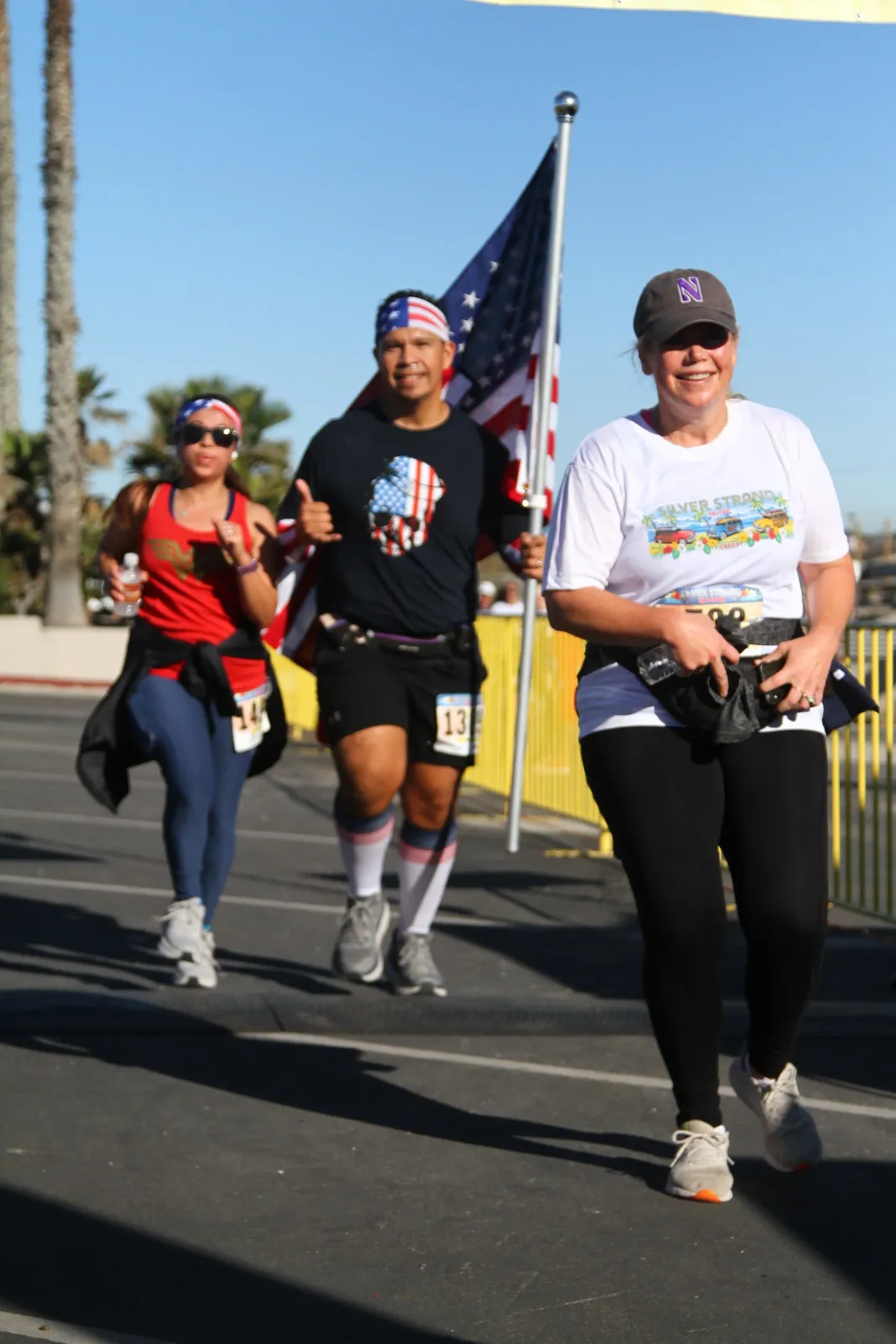 Three runners on a road course, one carrying an American flag, with palm trees in the background.