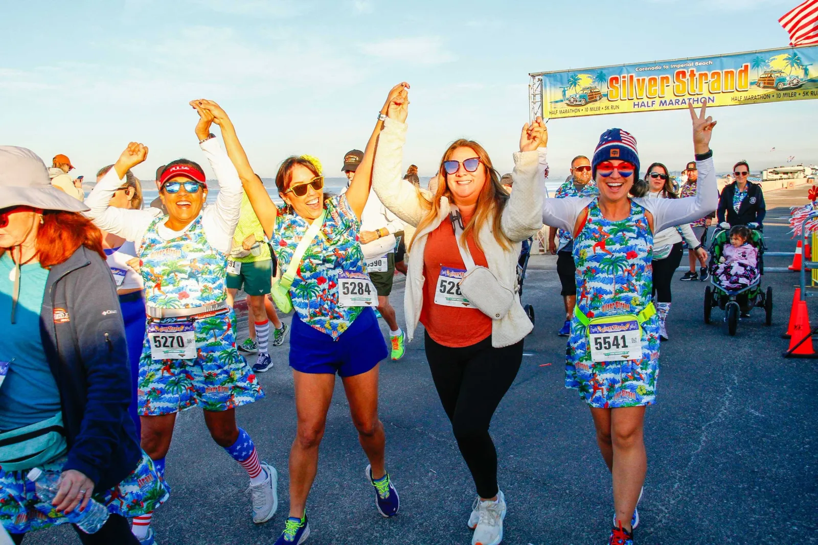Four women with their arms raised in celebration at the Silver Strand Half Marathon, with a race banner in the background.