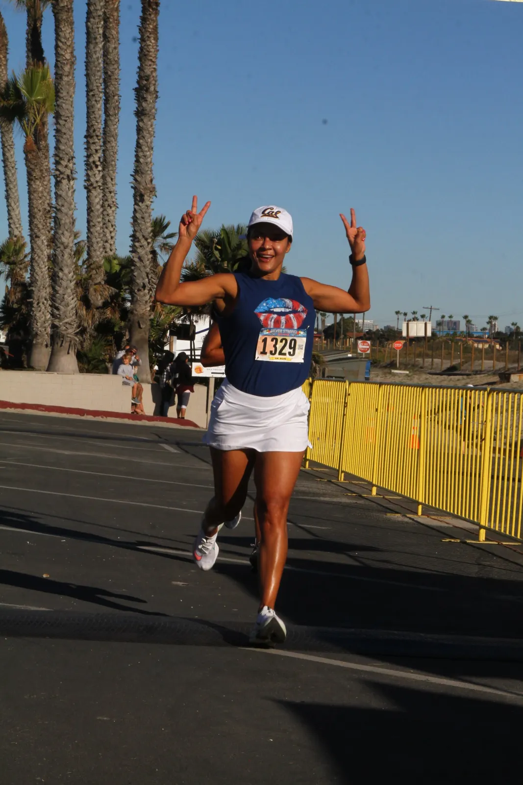 A female runner wearing a blue tank top with a patriotic lip design and a white skirt, smiles and holds up two peace signs while running on a road with palm trees in the background.