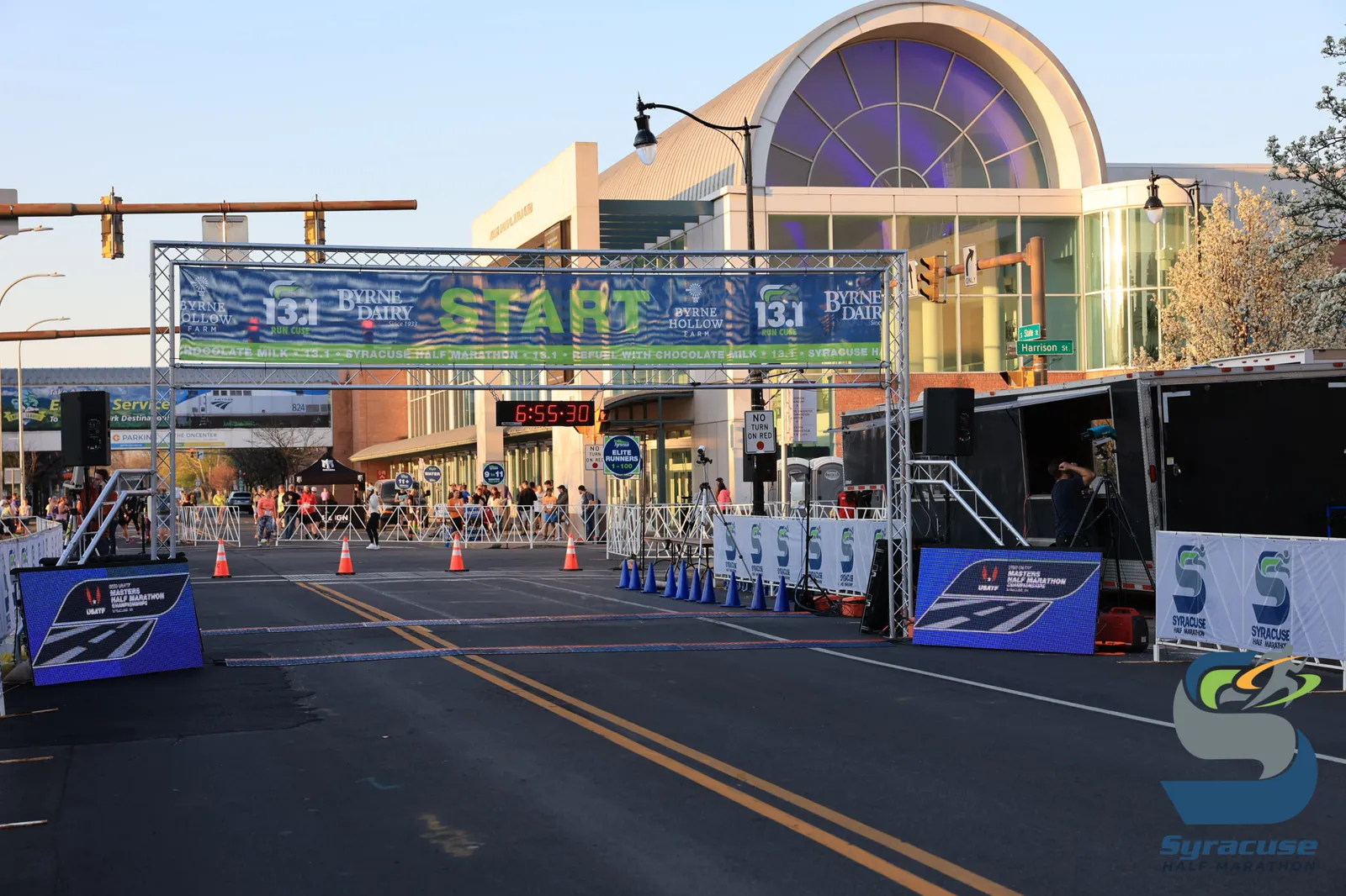 The start line of the Syracuse Half Marathon with a banner overhead and a crowd of runners gathered behind barriers.