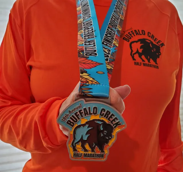 A person wearing an orange long-sleeved shirt holds up a Buffalo Creek Half Marathon medal.