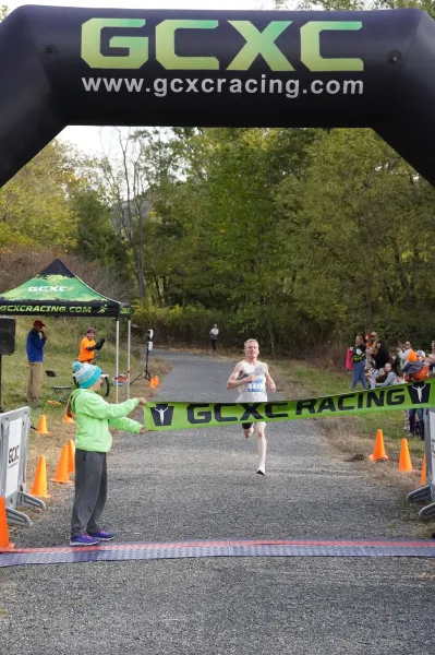 A male runner crosses the finish line, breaking a green banner that says GCXC RACING.
