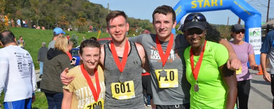 Four runners, two men and two women, pose for a photo with medals around their necks, smiling under a blue start/finish line arch.