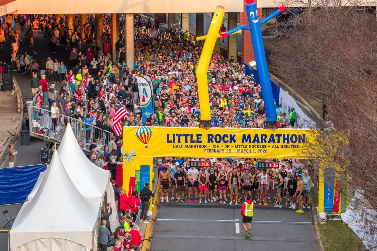 Runners gather at the start line of the Little Rock Marathon, with spectators cheering from the sidelines and inflatable figures flanking the banner.