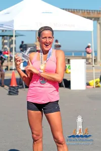 A smiling woman in a pink tank top and black shorts holds up a medal and a trophy after a race, with a tent and the ocean in the background.