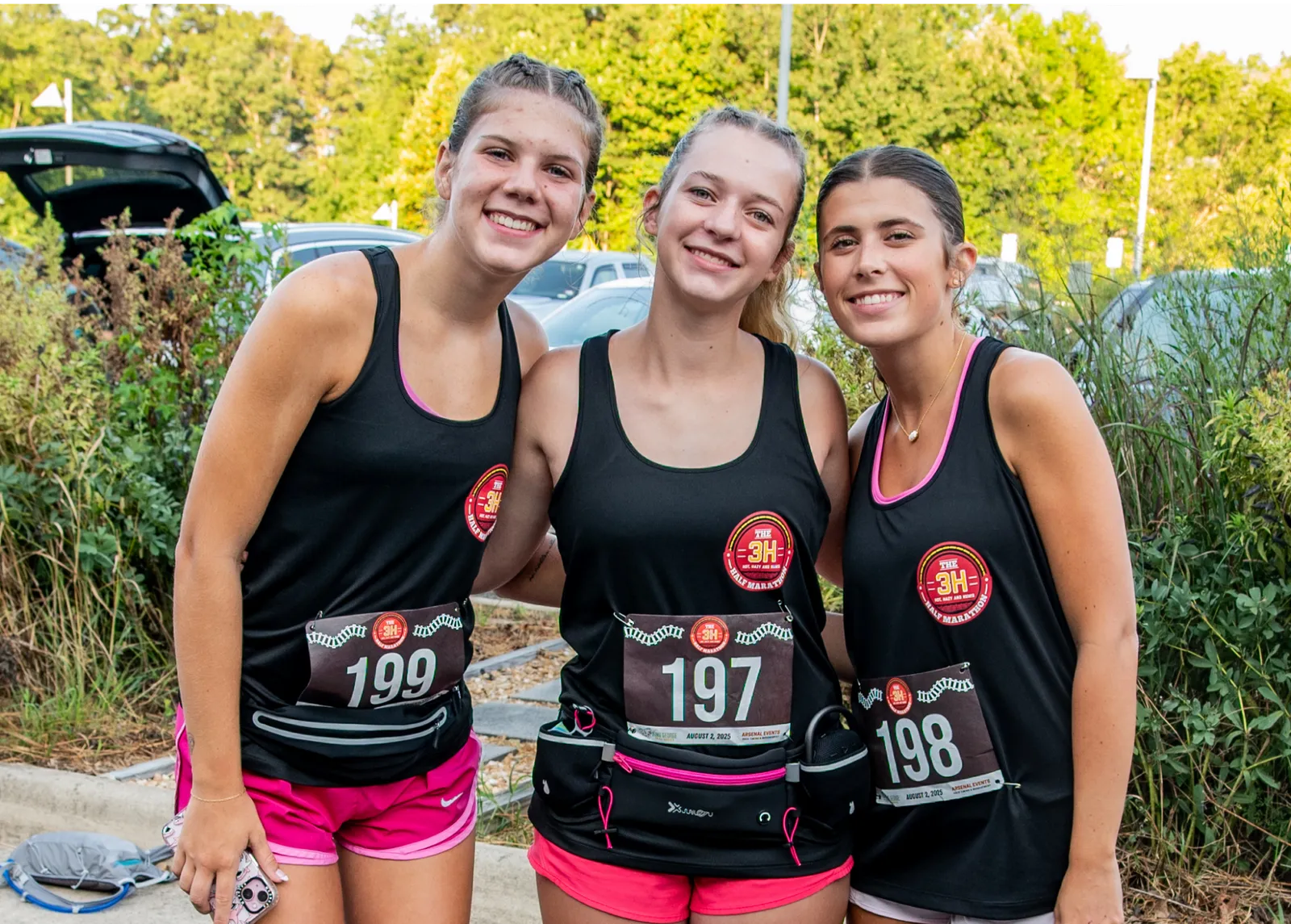 Three smiling young women wearing black tank tops with race bibs and pink shorts, posing for a photo before a race.