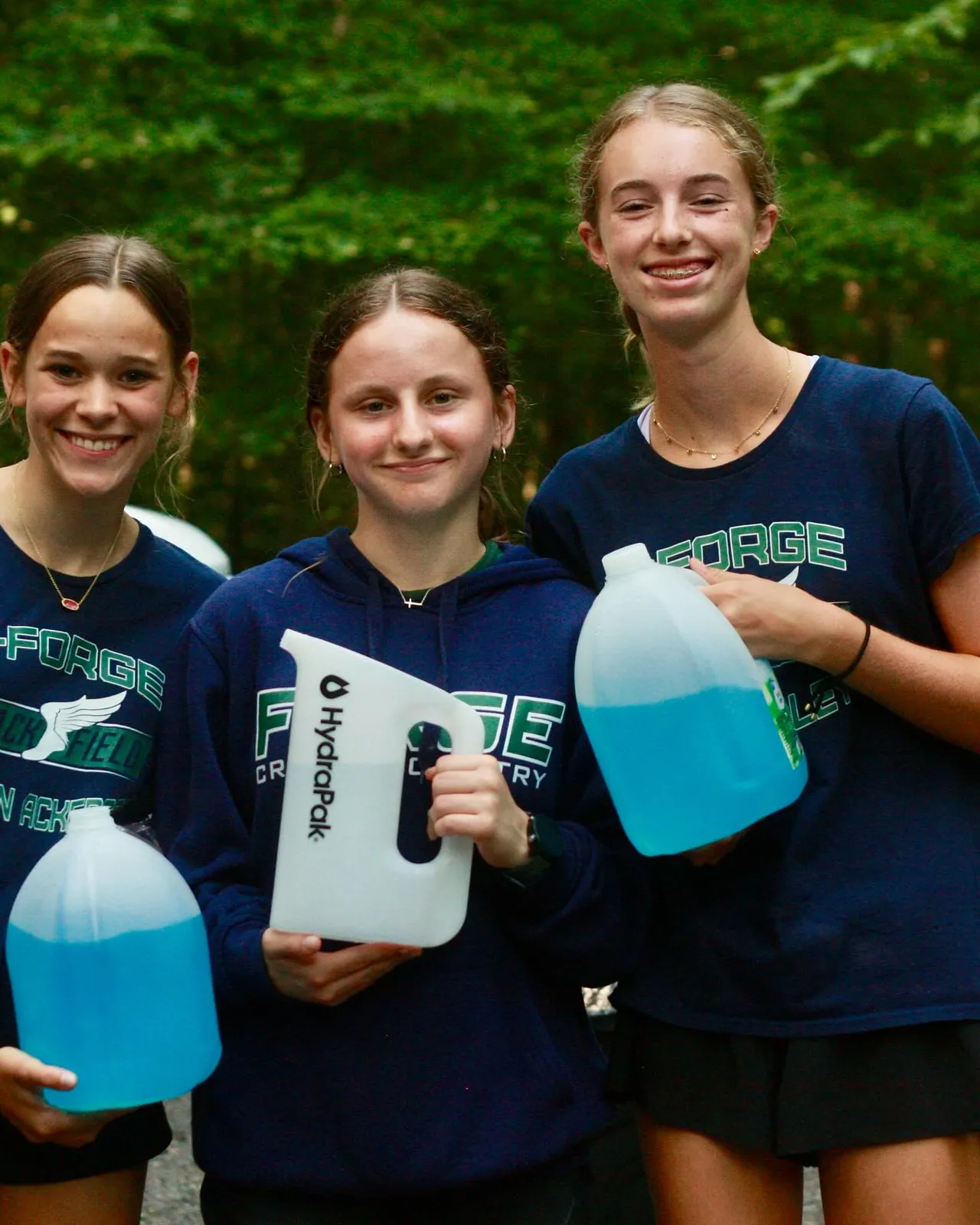Three young women smiling and holding water jugs at a race event.