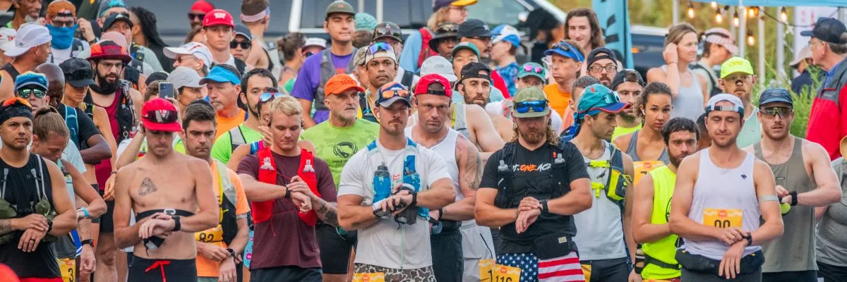 A diverse group of runners in athletic wear stand at the start line, many checking their watches, anticipating the start of a race.