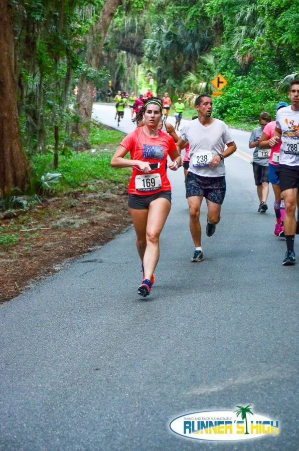 A female runner with bib number 169 in a red shirt runs on a tree-lined road, with other runners behind her.