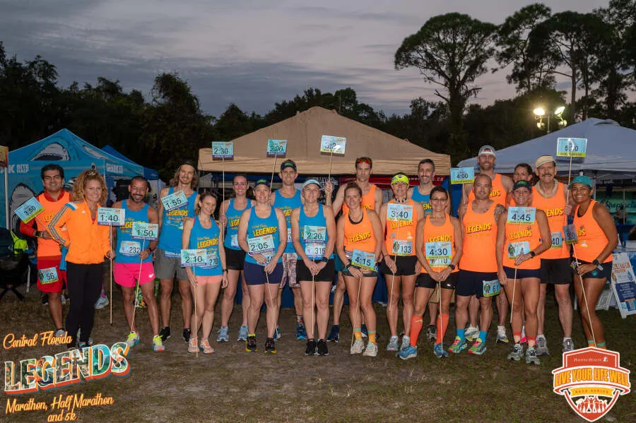 A group of runners in orange and blue shirts pose for a photo at the Central Florida Legends Marathon, Half Marathon, and 5K, holding signs with pace times.