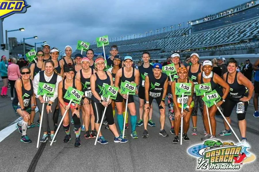 A group of runners pose for a photo holding pace signs at the start of a race, with a stadium in the background.