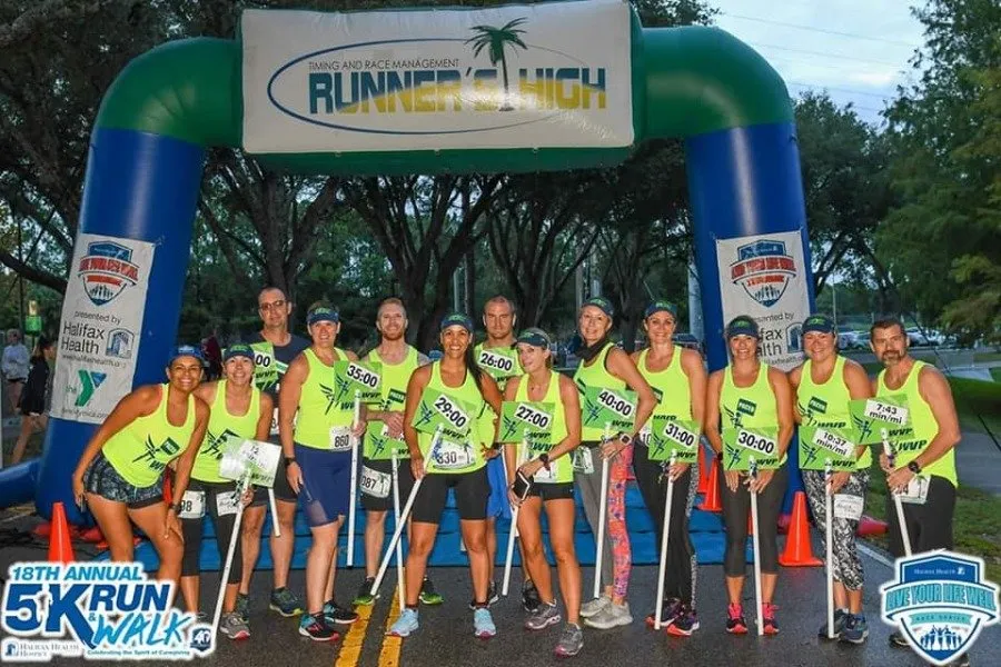 A group of runners in neon green shirts stand under an inflatable archway at the start or finish line of a race, holding signs with times.