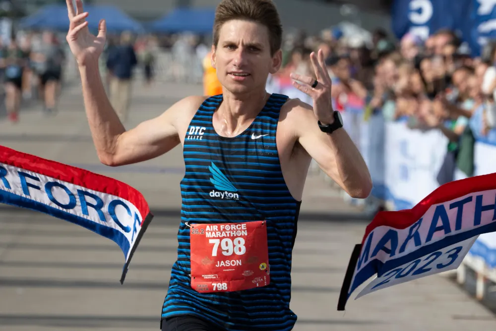 A male runner, Jason, wearing a blue and black striped tank top with bib number 798, crosses the finish line of the Air Force Marathon 2023 with his arms raised in celebration, with spectators in the background.