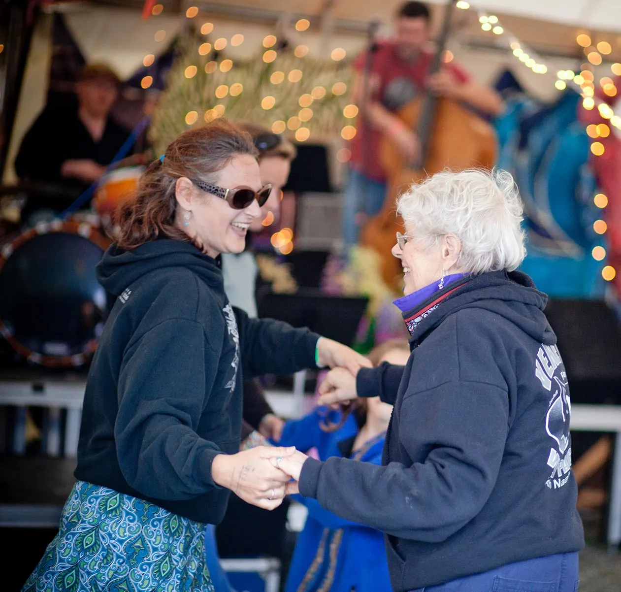 Two women smiling and holding hands while dancing, with a band playing in the background.