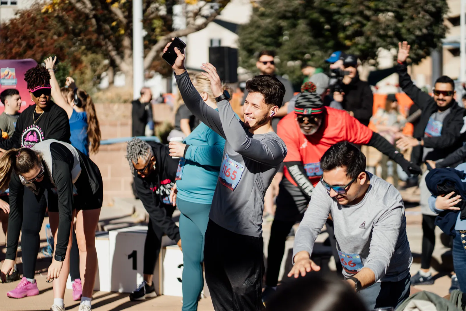 A group of runners are gathered at the start line of a race, with some stretching and others taking photos with their phones. The atmosphere appears energetic and excited.