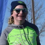 A runner wearing sunglasses and a beanie smiles at the camera, wearing a green and grey hoodie with text and a mountain graphic.