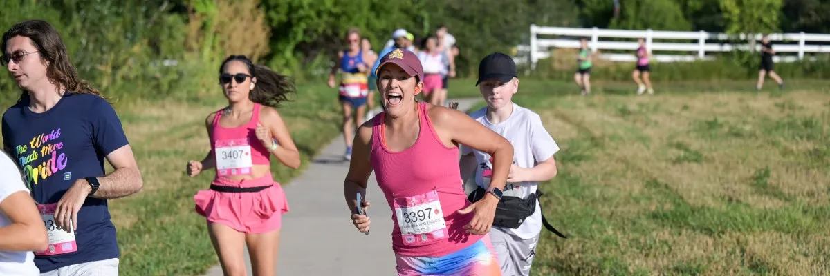 A group of runners, including a woman with an excited expression in the foreground, run along a paved path with grassy areas and trees in the background.