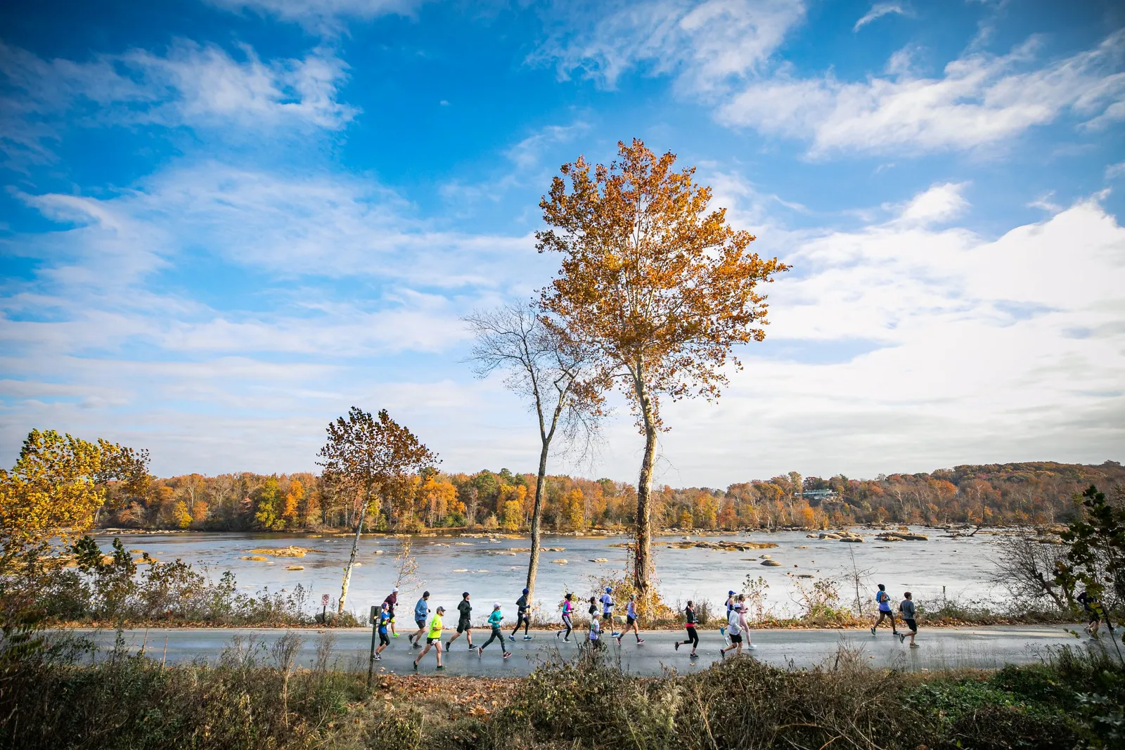 Runners race along a road next to a river with fall foliage on the banks under a blue sky with clouds.