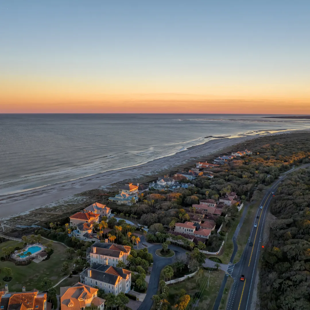 Aerial view of Amelia Island coastline at sunset, showing the beach, ocean, a road, and residential buildings.