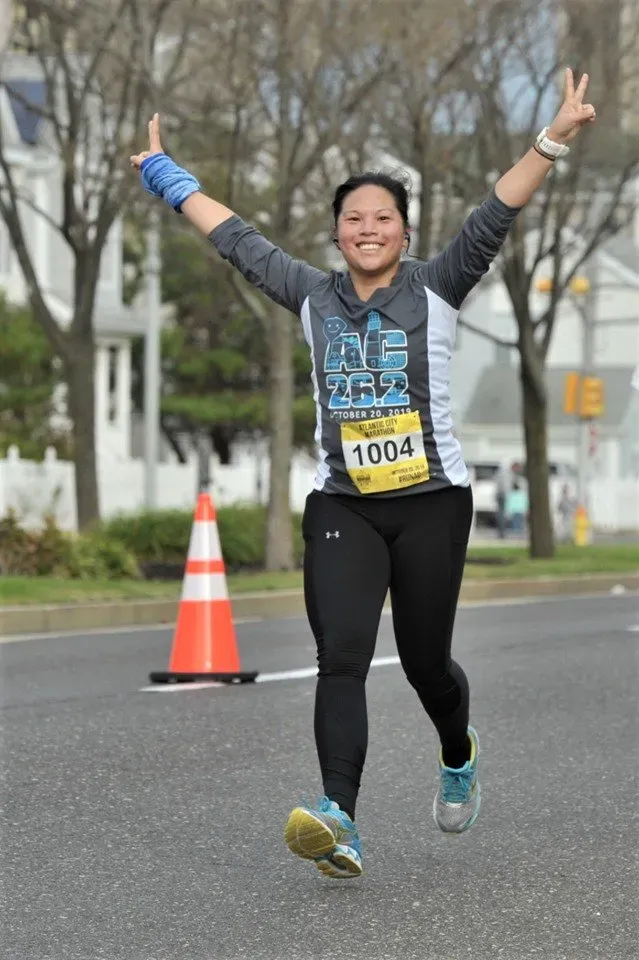 A female runner with arms raised in celebration, wearing a race bib and smiling.