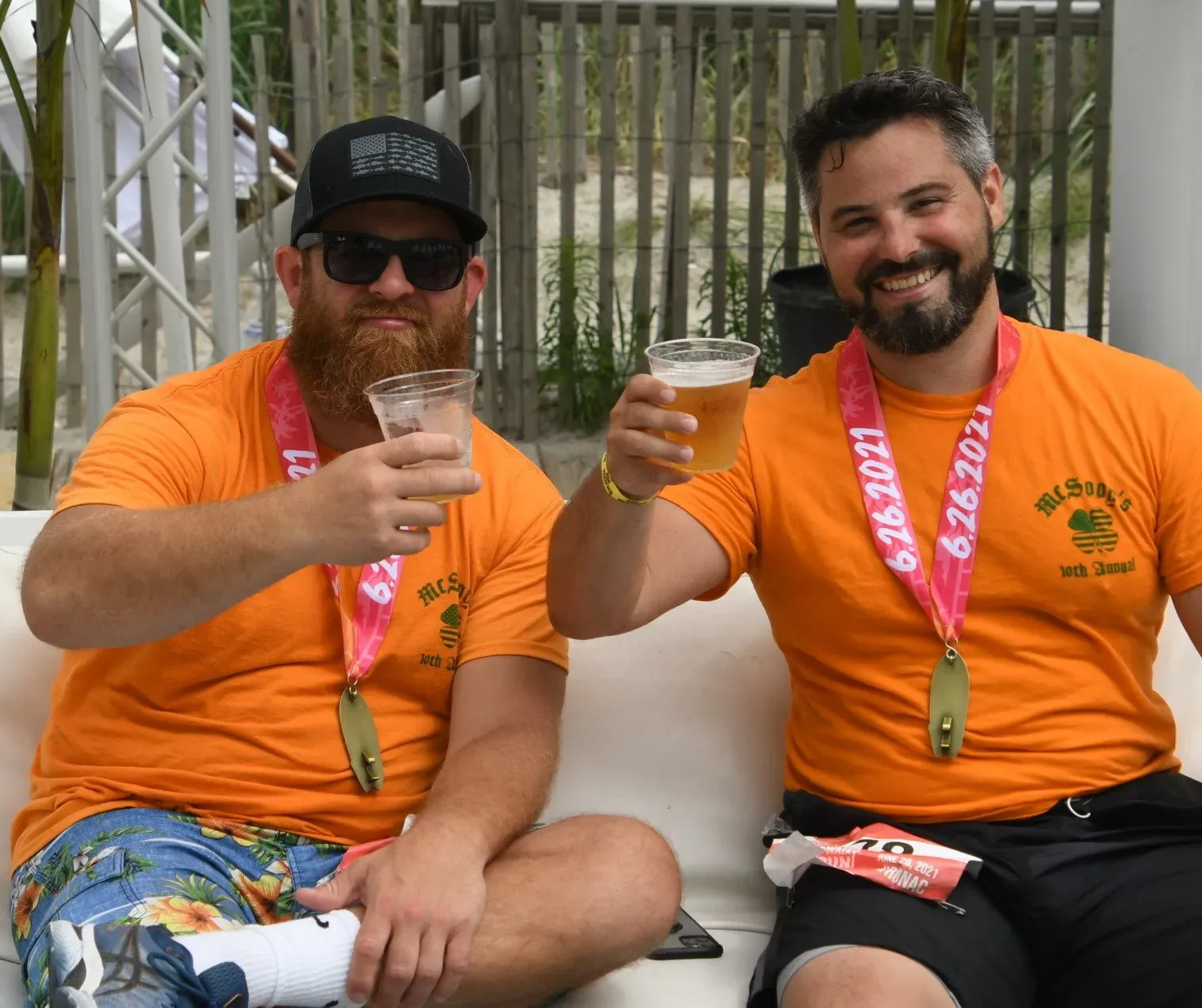 Two men in orange t-shirts and race medals are sitting and toasting with beers.