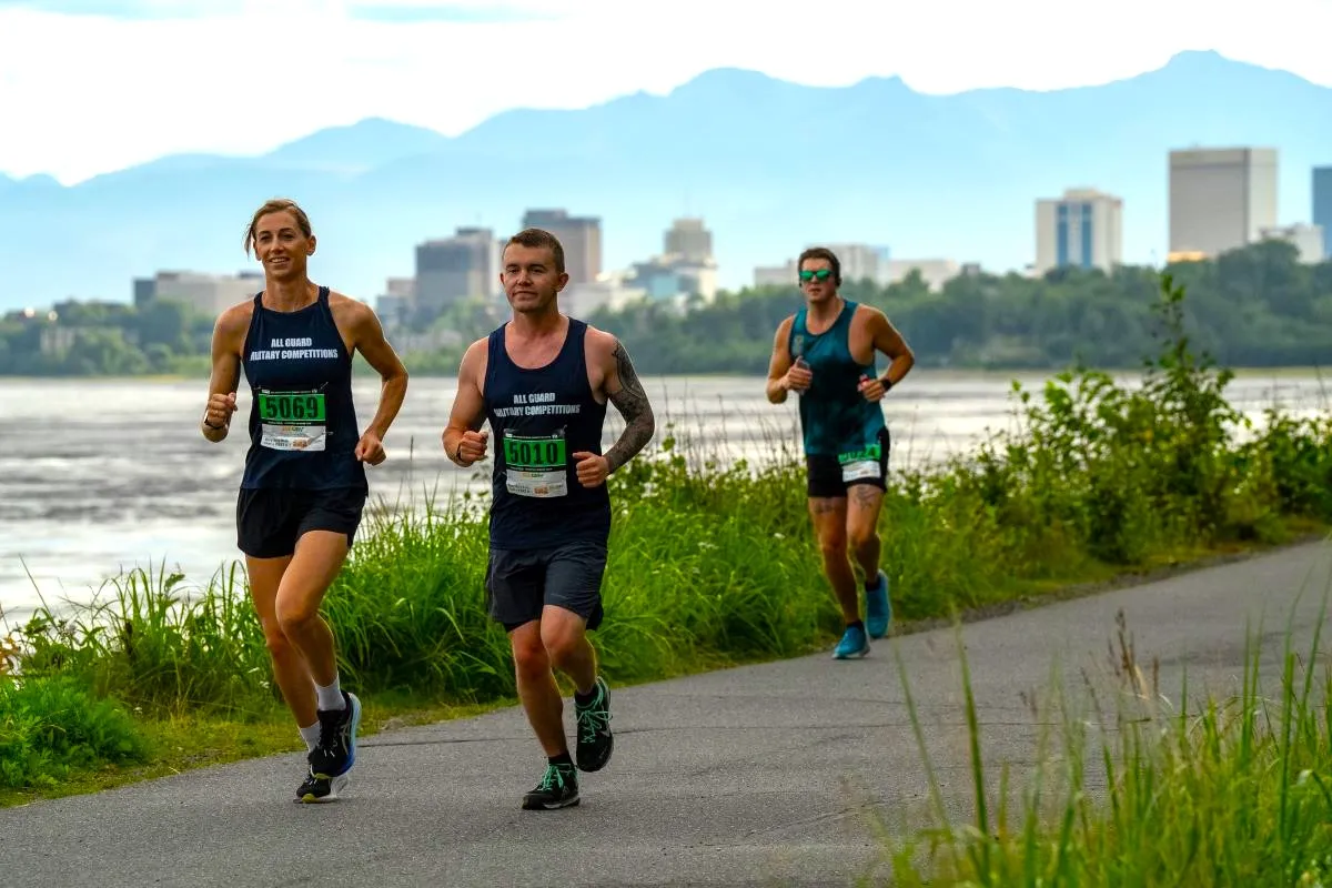 Three runners on a paved path with a body of water to their left and a city skyline and mountains in the background.