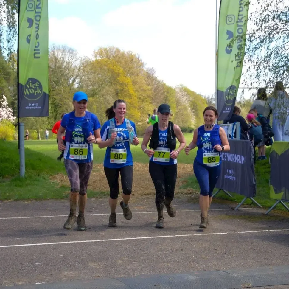Four female runners in blue race shirts with bib numbers 86, 80, 77, and 85 running towards the camera on a paved path, with green banners and trees in the background.