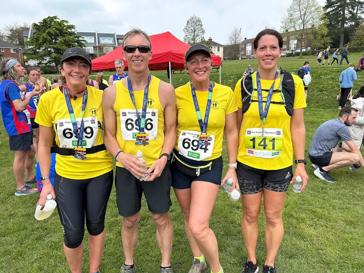Four runners in yellow shirts and medals smiling after a race, with a red tent and other participants in the background.