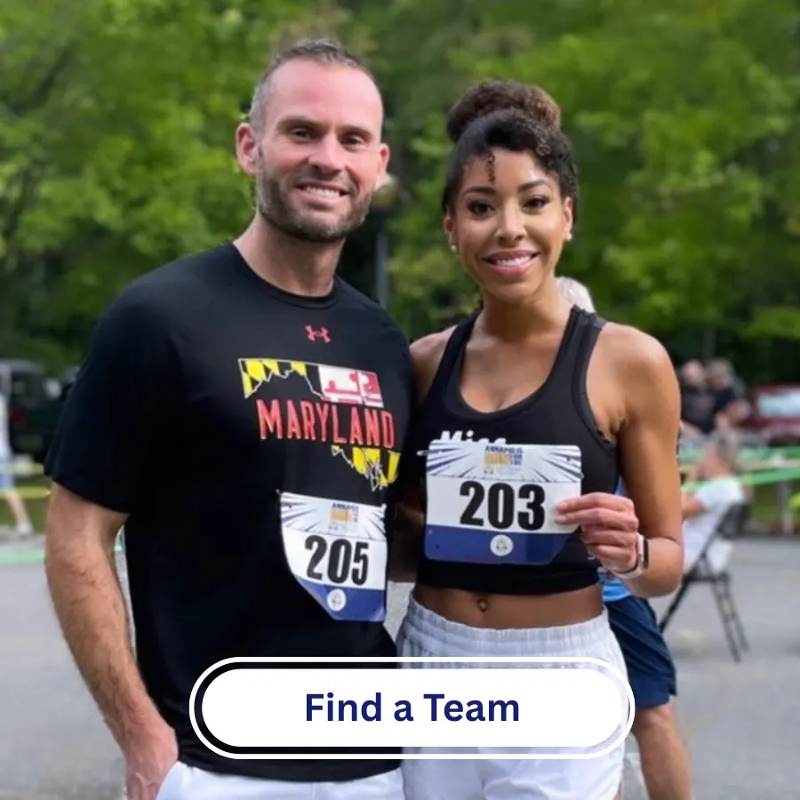 A man and a woman, both wearing race bibs, smile at the camera. The man wears a black t-shirt with 'Maryland' on it, and the woman holds up her bib.