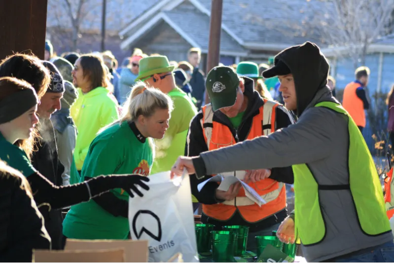 People in green shirts and high-visibility vests interacting at a table with bags and cups, likely at the start of a race.